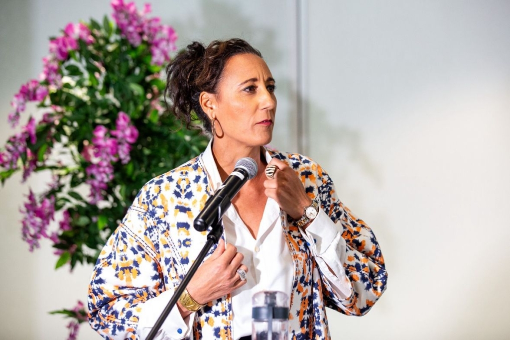 Michelle Moore, a Black woman with light skin, stands in front of a microphone wearing a blue and yellow colourful jacket over a white shirt. She stands strong and emboldened. 