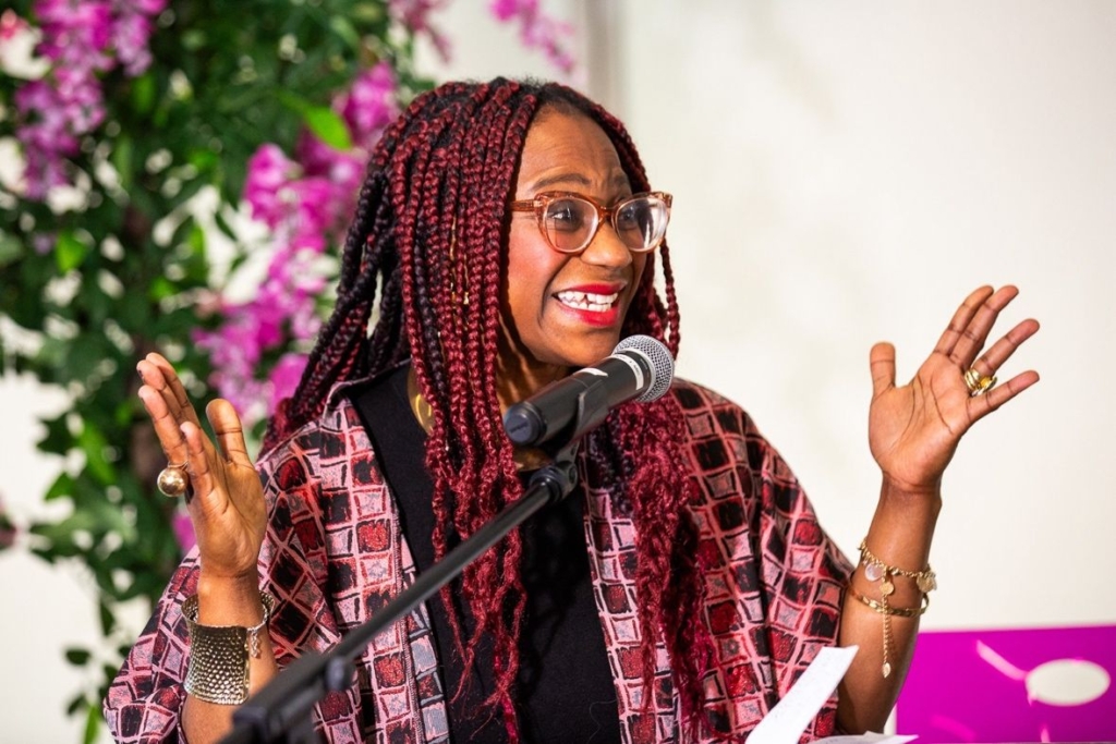 A Black woman, Peju Abuchi, stands infront of a microphone with her hands expressively either side of her face. She wears red lipstick and a huge infectious smile. 