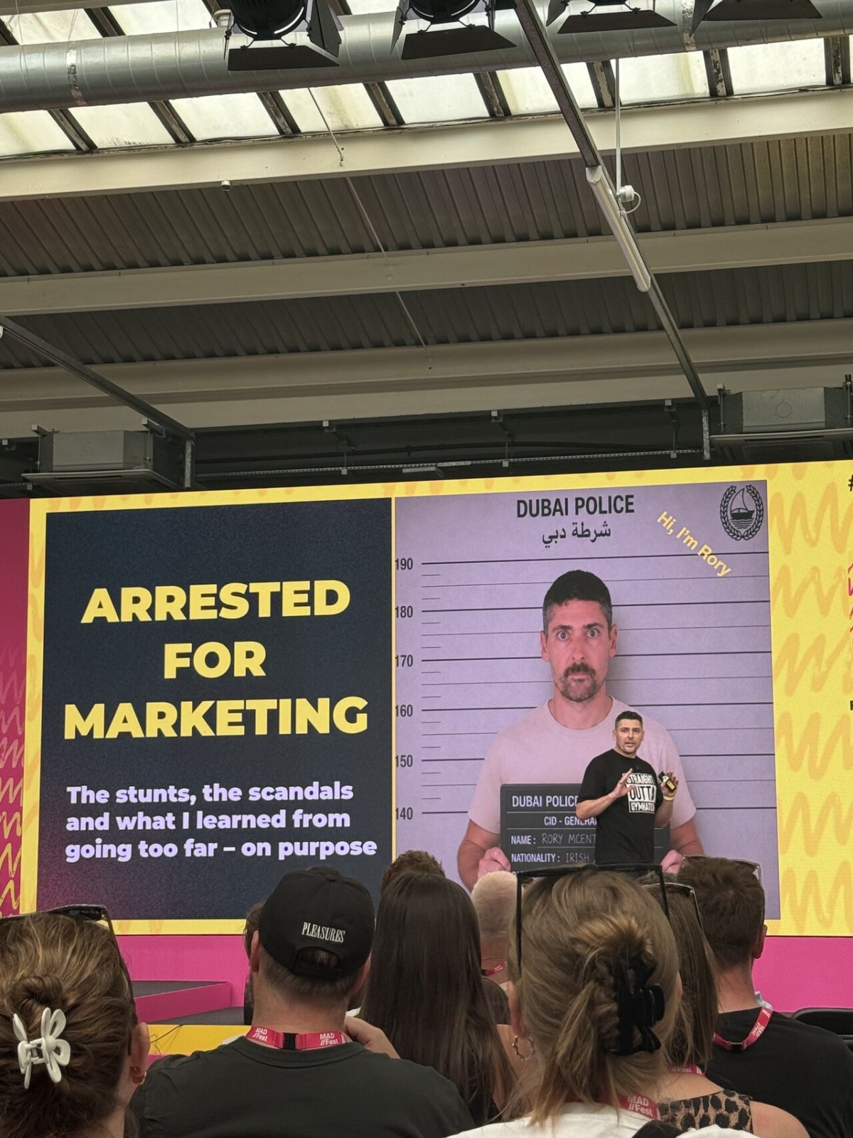 A man addresses an audience at a conference, with a sign that says "Arrested for Marketing" in the background.