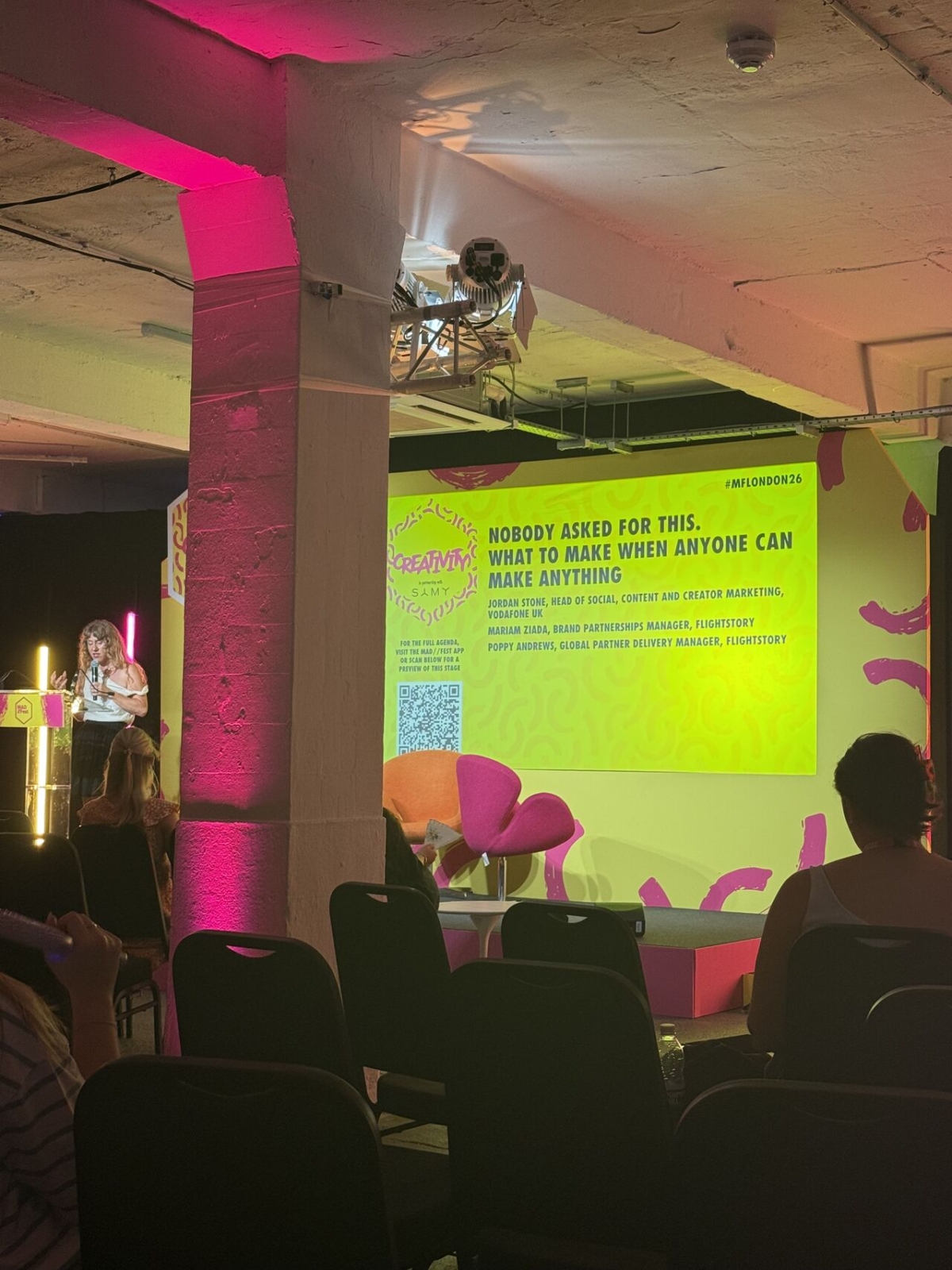 A woman speaks to an audience, pointing at a large screen that showcases her presentation: "Nobody asked for this. What to make when anyone can make anything".