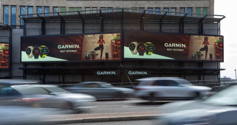 Garmin smartwatch ads span a city building facade as cars blur past in traffic, creating a fast-paced urban scene with a modern, energetic tone.