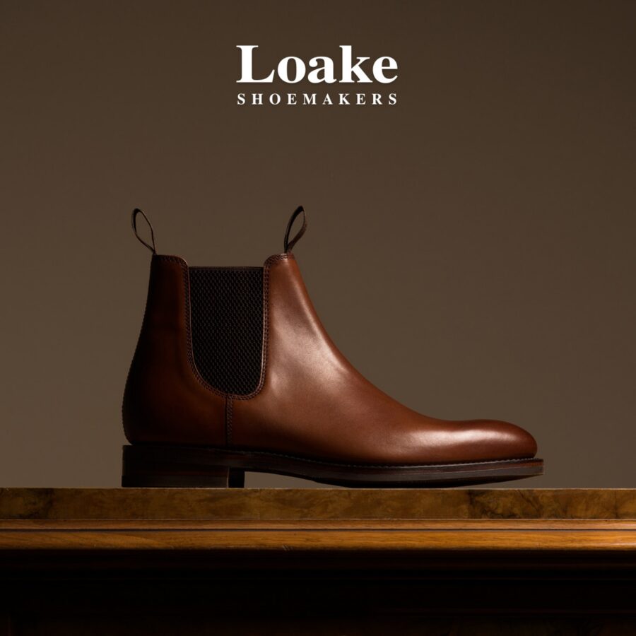 A polished brown leather Loake Chelsea boot with black elastic sides rests on a wooden shelf against a dark brown backdrop, conveying classic craftsmanship.