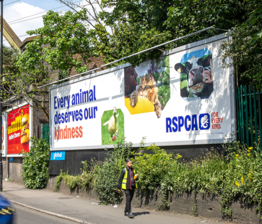 A large RSPCA OOH billboard reads “Every animal deserves our kindness,” with animal photos above a worker walking past greenery on a sunny street.