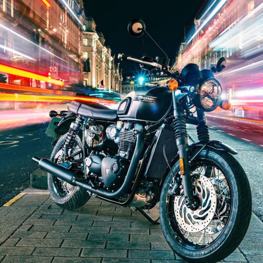 A black Triumph motorcycle on a city street at night as colorful traffic trails streak past historic buildings, creating a dramatic, fast-paced cinematic mood.