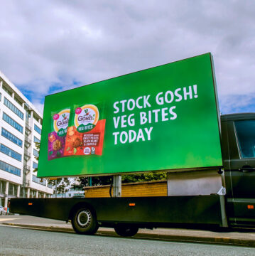 A bright green billboard on a truck advertises “Stock Gosh! Veg Bites Today,” with product packaging pictured; urban buildings and cloudy skies frame the lively street scene.