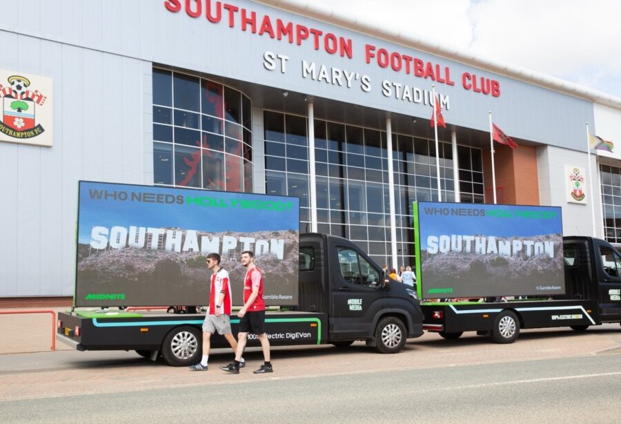Two Southampton football fans walk past promotional digivans outside St Mary’s Stadium, beneath the club sign, with the Midnite ads showing Southampton in hollywood-esque letters on the screen.