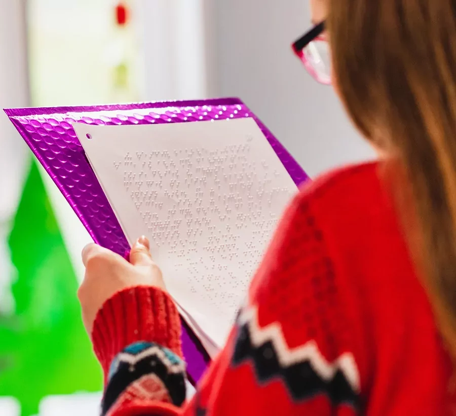 A girl in glasses and a red patterned sweater reads a braille page on a purple clipboard, with a bright green Christmas tree blurred in the background.