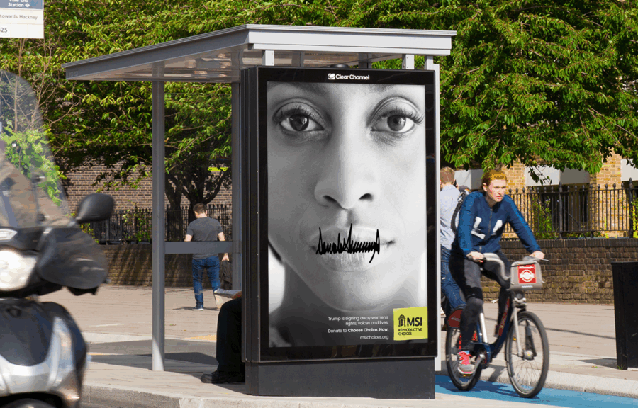 A grayscale close-up of a woman’s face on a bus-stop poster, with Trump's angry signature written across her mouth, silencing her. Cyclists pass in bright daylight; stark, urgent tone. This is the OOH print ad from MSI's Signed Away campaign.