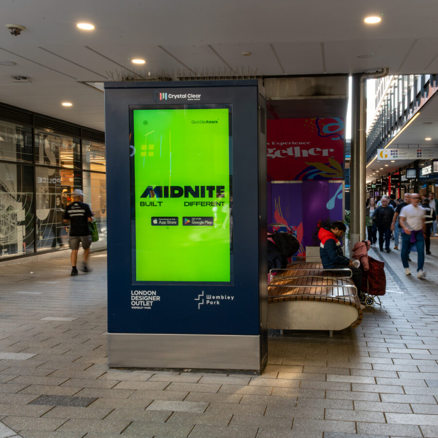 A bright green Midnite digital ad stands in a busy indoor shopping arcade, as shoppers pass storefronts and people sit on a nearby bench.