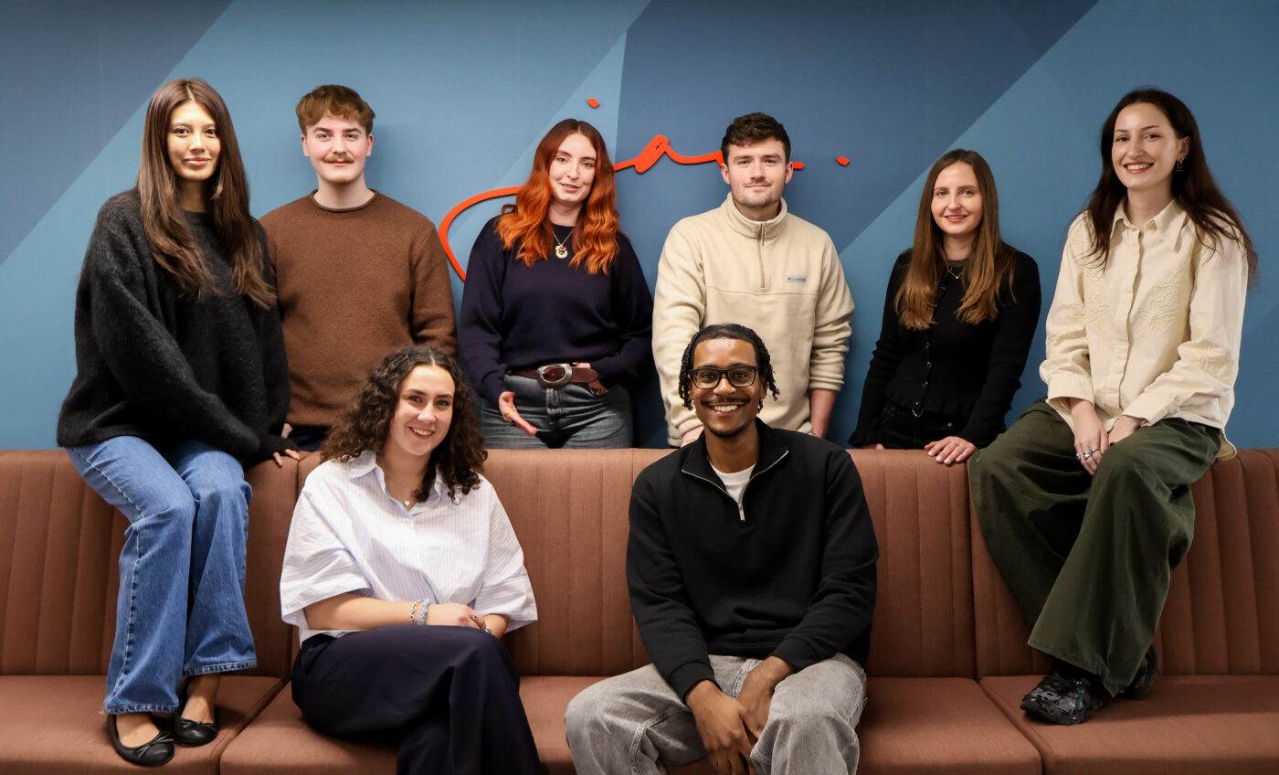 Team of work colleagues are gathers with backs to a wall and two sitting on a sofa