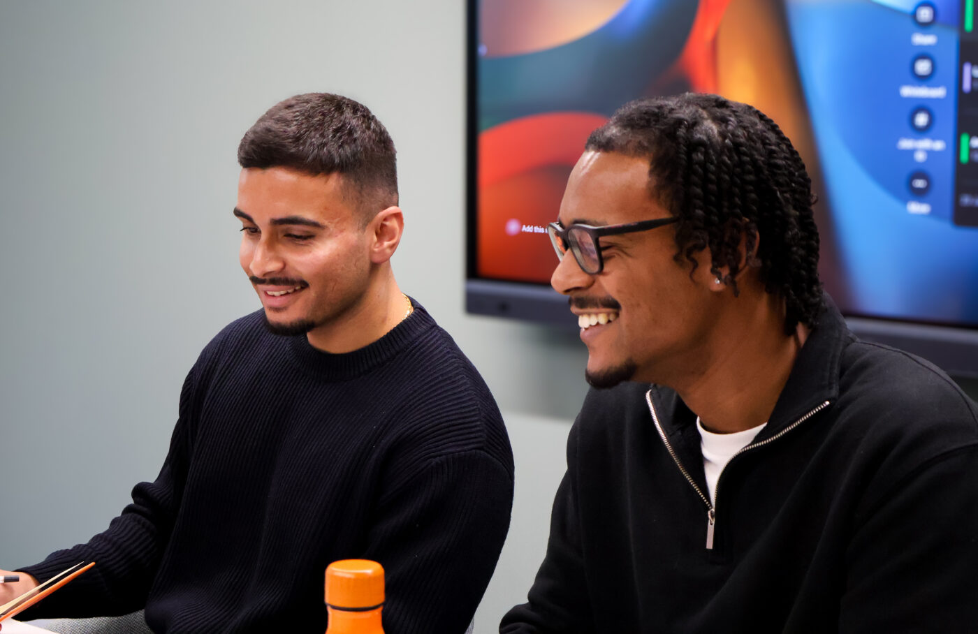 Two co-workers smile in a meeting with a screen behind them