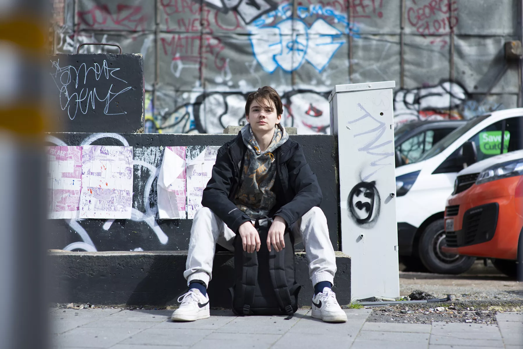 A young person sits on a black suitcase before a graffiti-covered wall, with parked cars nearby; the urban scene feels gritty and contemplative.