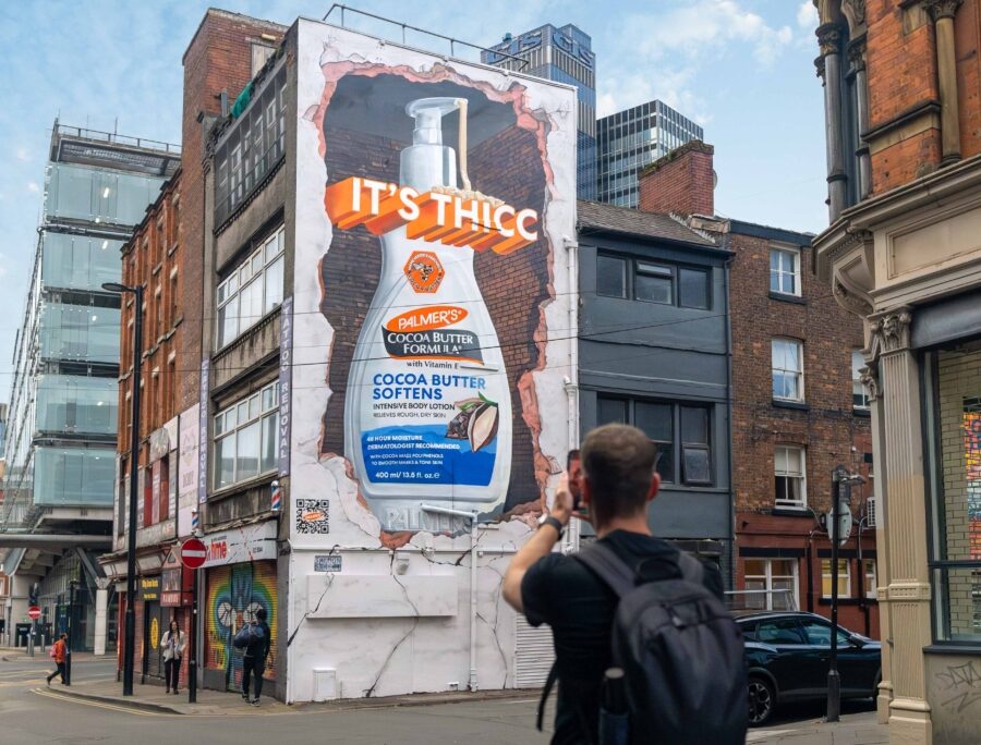 A person in the foreground takes a photo of a large mural on a brick building depicting a bottle of Palmer’s Cocoa Butter Formula lotion with the text “IT’S THICC” above it.