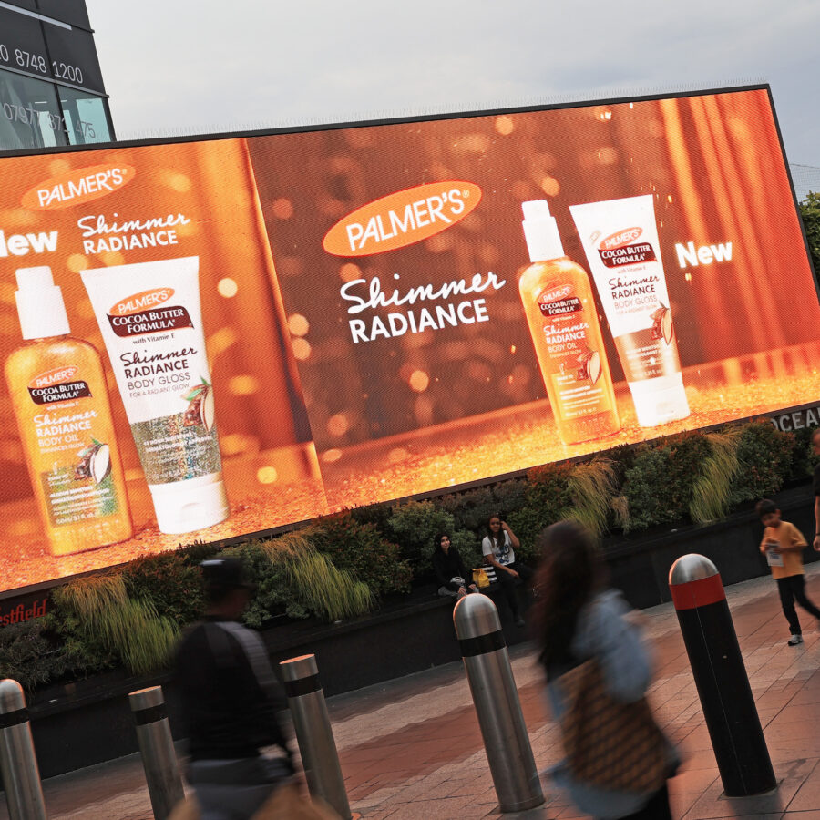 A large digital billboard advertising Palmer’s Cocoa Butter Formula Shimmer Radiance body oil and body gloss, positioned above the pavement at Westfield White City, with pedestrians walking underneath.