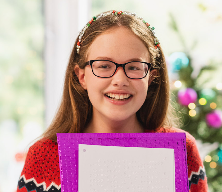 A smiling girl (the ad’s star, Keira, who is blind) wearing glasses and a festive headband holds a purple-bordered certificate in front of a decorated Christmas tree.