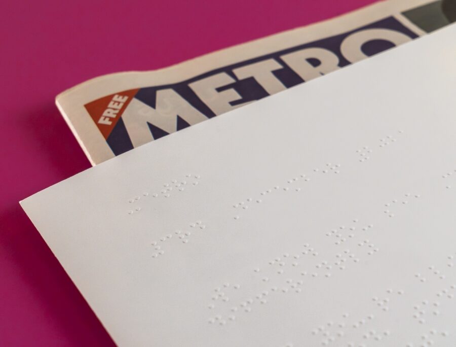 A white Braille-embossed sheet covers most of a Metro newspaper, leaving its masthead visible against a vivid pink background, suggesting accessible news.