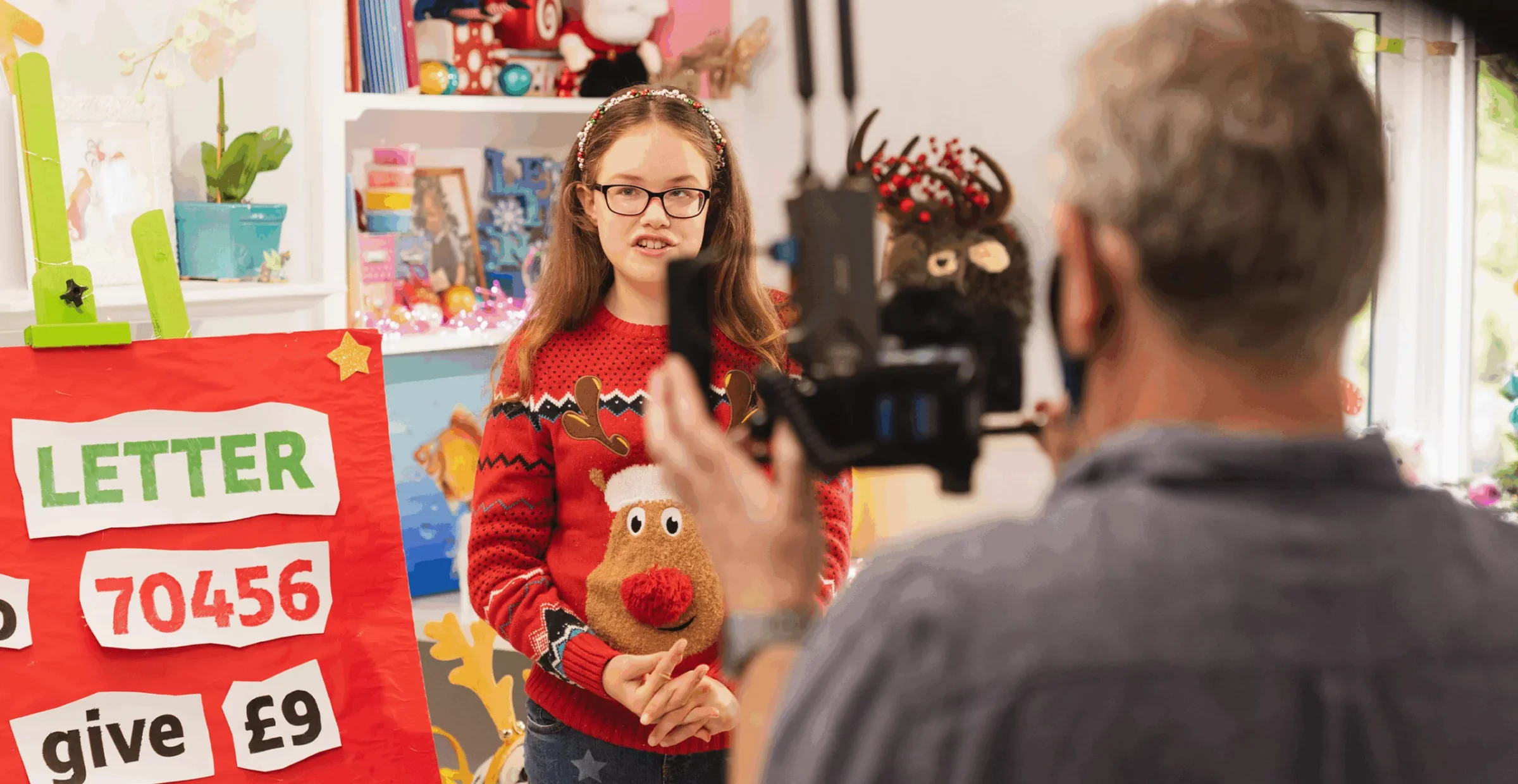 A smiling girl in a red reindeer sweater speaks to a camera beside a sign reading “LETTER 70456, give £9”; cheerful interview setting.