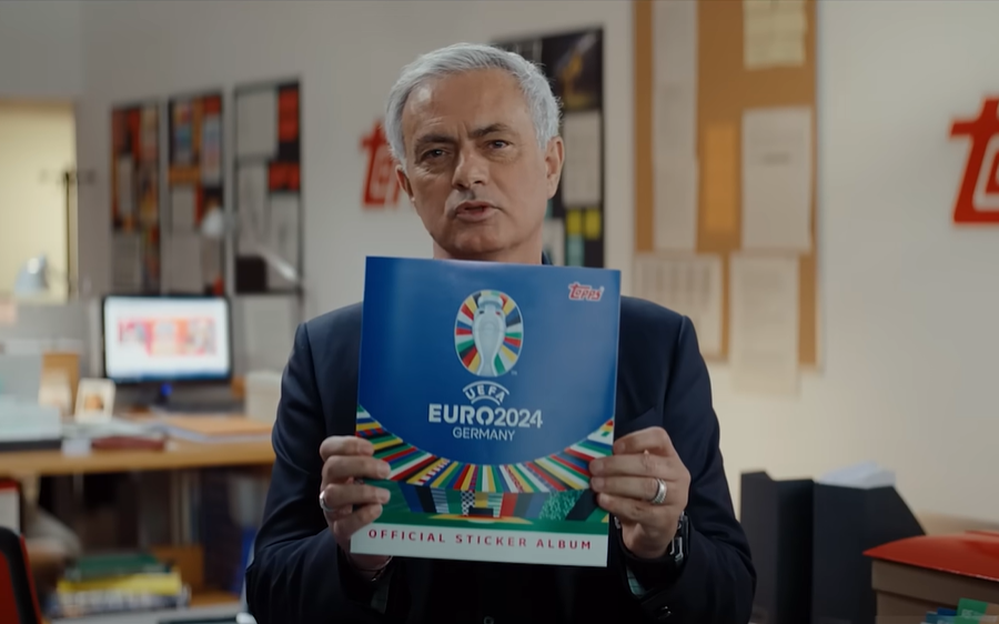 Jose Mourinho presents a blue UEFA EURO 2024 official sticker album in a Topps office, looking serious and confident amid desks and posters.