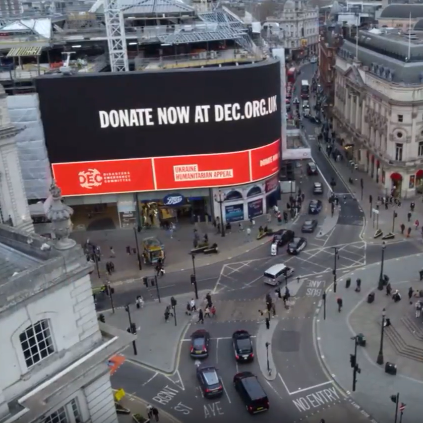 Aerial view of a busy Piccadilly Circus beneath a giant curved billboard reading “Donate now at DEC.org.uk,” promoting a humanitarian appeal amid traffic and pedestrians.