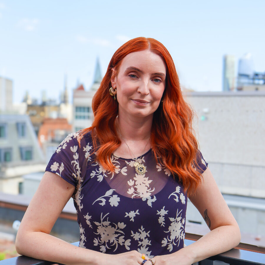 A red headed women looks down the lens of the camera with a blue sky behind her