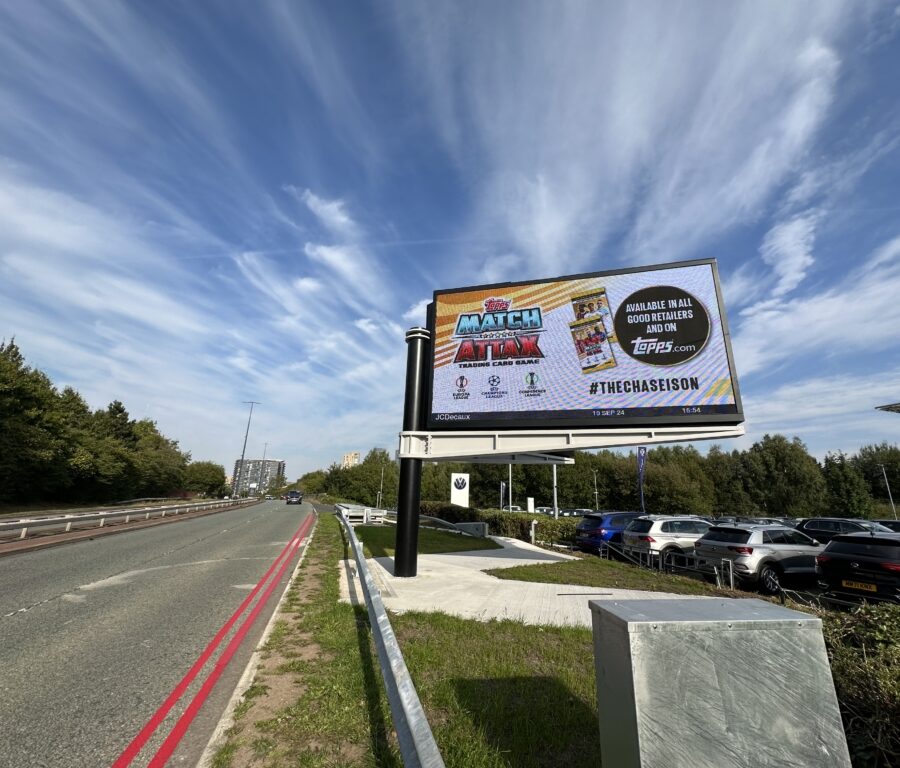 A roadside billboard advertises Topps Match Attax football cards beside a busy road and parked cars, beneath dramatic streaked clouds; the scene feels expansive and energetic.