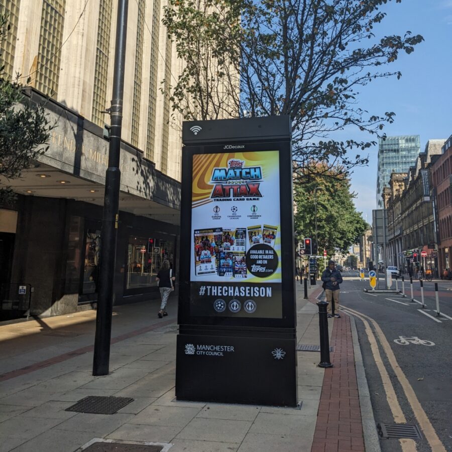 A Manchester City Council digital street display billboard advertises a “Match Attack” ad, framed by tall city buildings and leafy trees on a sunny urban street.