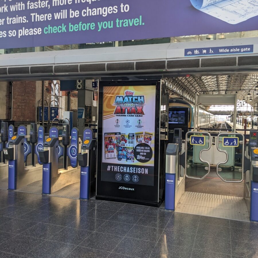 A train station’s wide-aisle gate area with blue ticket barriers and accessible-gate signs, beside a Match Attax advertisement; shown on a vertical digital billboard.