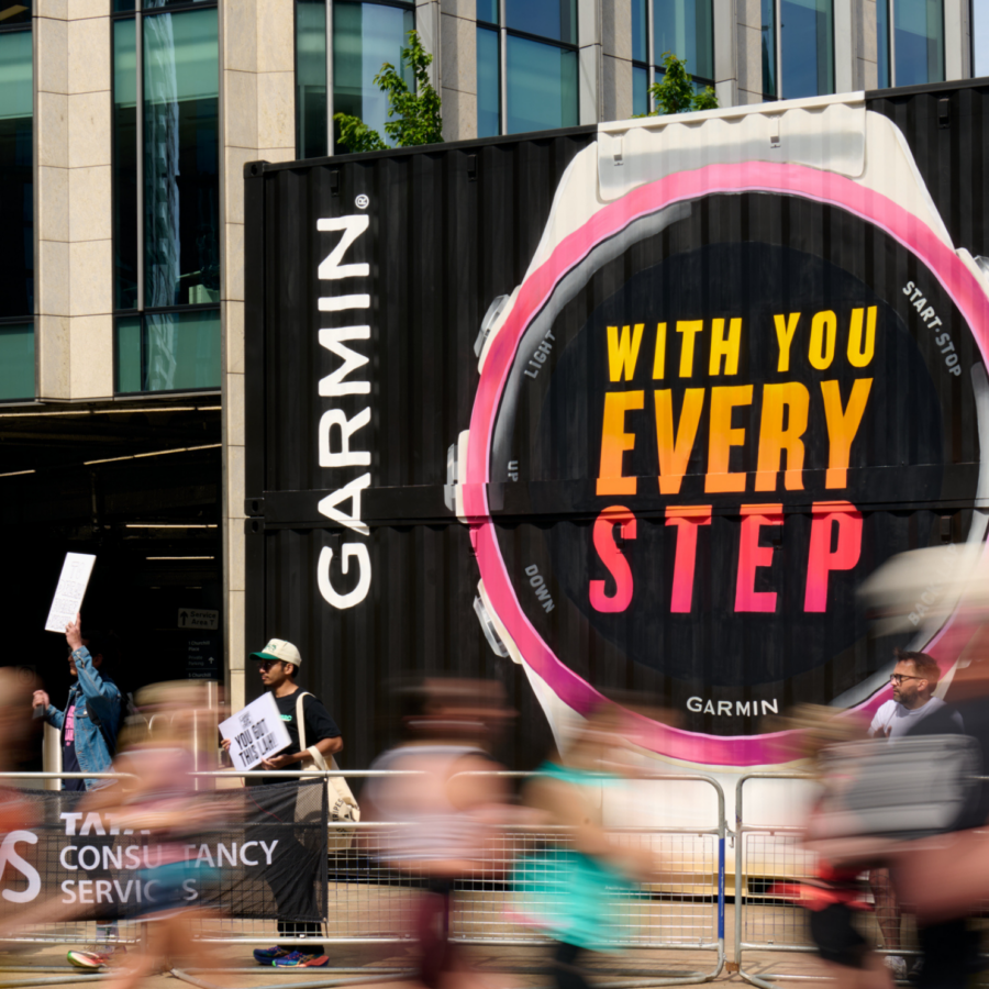 Blurred runners race past a large Garmin watch display reading “With you every step” on a city street, conveying energetic support for runners.