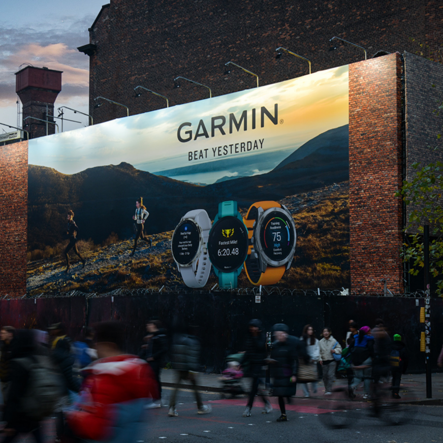 A giant Garmin billboard in central Manchester, showing sports watches and a mountain runner towers over a busy city street, where blurred pedestrians pass at dusk. The mood is energetic.