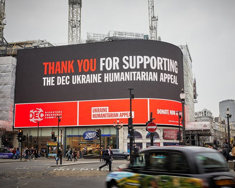 A giant billboard on Piccadilly Circus reads “Thank you for supporting the Ukrainian humanitarian appeal” above a busy street, with pedestrians, traffic, and construction cranes.