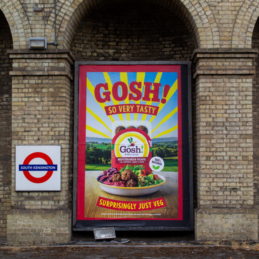 A bright red-framed Gosh! vegan food poster stands in a brick railway arch at South Kensington tube station, showing a colourful vegetable dish beneath bold “So Very Tasty” text.
